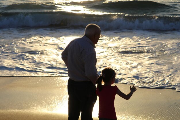 Foto vista traseira de uma menina com o avô de pé na costa na praia