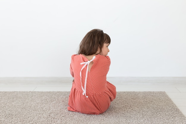 Vista traseira de uma menina em um vestido de bolinhas vermelhas, sentada no chão em uma parede branca.