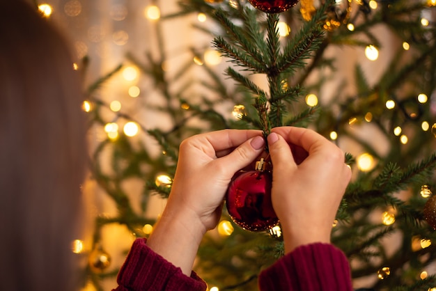 Vista traseira de uma menina morena com suéter cor de vinho, pendurando uma bola vermelha na árvore de Natal