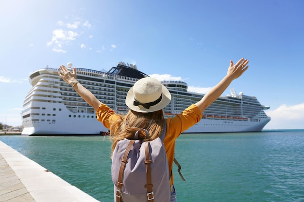 Foto vista traseira de uma menina viajante com os braços levantados em frente a um grande navio de cruzeiro