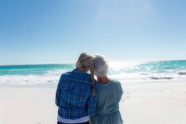 Vista traseira de uma mulher caucasiana e sua mãe de pé na praia com o céu azul e o mar ao fundo, abraçando e olhando para longe