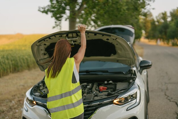 Vista traseira de uma mulher com um colete refletivo abrindo o capô de seu carro