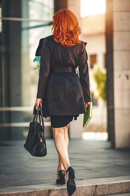 Vista traseira de uma mulher de negócios está andando no caminho no prédio de escritórios. Ela está segurando uma bolsa e indo para o trabalho.