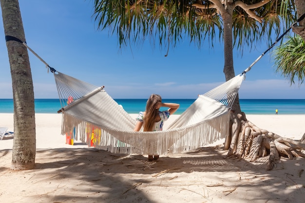 Vista traseira de uma mulher de viagens sentada em uma rede branca na praia de areia