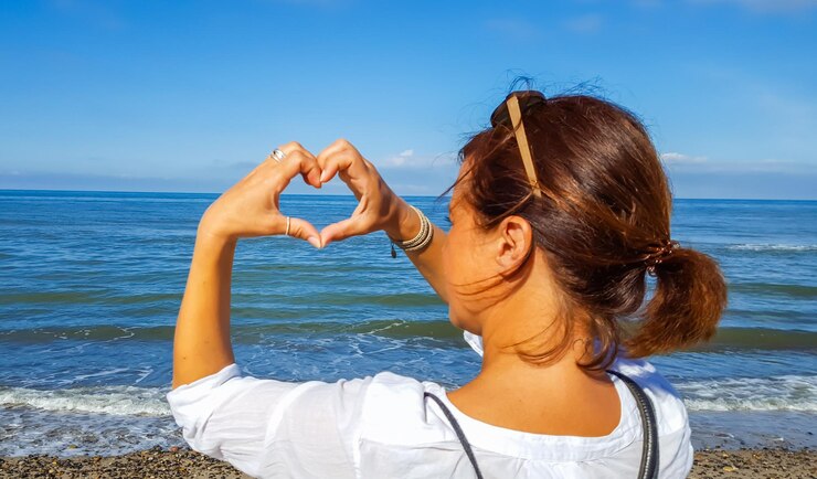 Foto vista traseira de uma mulher fazendo forma de coração com as mãos contra o mar