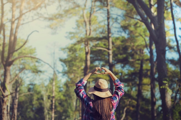 Vista traseira de uma mulher fazendo forma de coração sobre a cabeça enquanto está de pé contra árvores na floresta