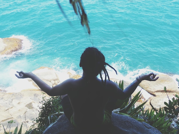 Vista traseira de uma mulher fazendo ioga em um penhasco ao lado do mar