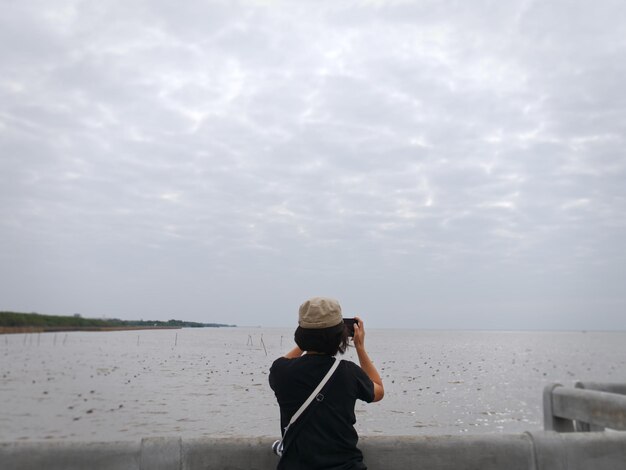 Foto vista traseira de uma mulher fotografando o mar contra o céu