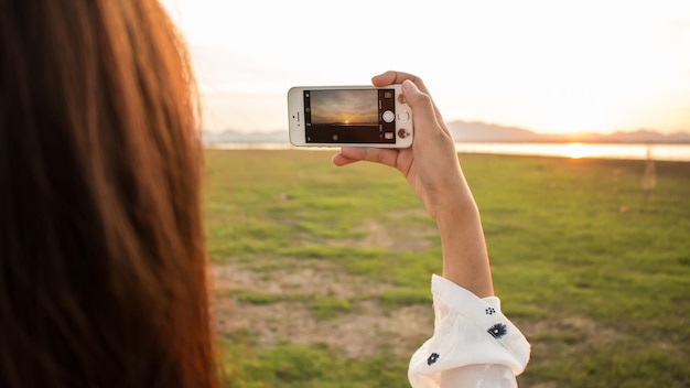 Foto vista traseira de uma mulher fotografando um lago através de um telefone inteligente ao pôr do sol