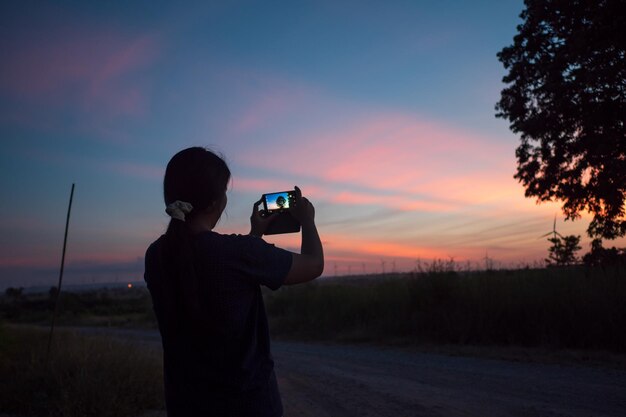 Foto vista traseira de uma mulher fotografando uma árvore através de um telefone móvel durante o pôr do sol