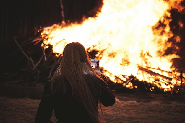 Foto vista traseira de uma mulher fotografando uma fogueira com um telefone celular à noite