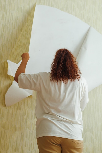 Vista traseira de uma mulher gorda vestindo calças bege de camiseta branca arrancando o papel de parede amarelo da parede Vertical