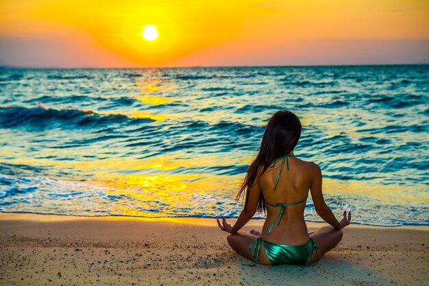 Vista traseira de uma mulher mediando na praia durante o pôr-do-sol