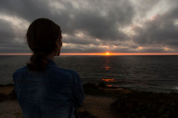 Foto vista traseira de uma mulher olhando para a costa do mar contra o céu ao pôr do sol