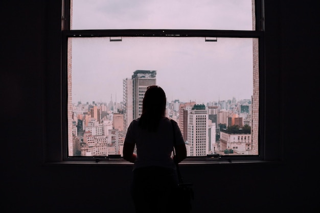 Vista traseira de uma mulher olhando para a paisagem da cidade através de uma janela