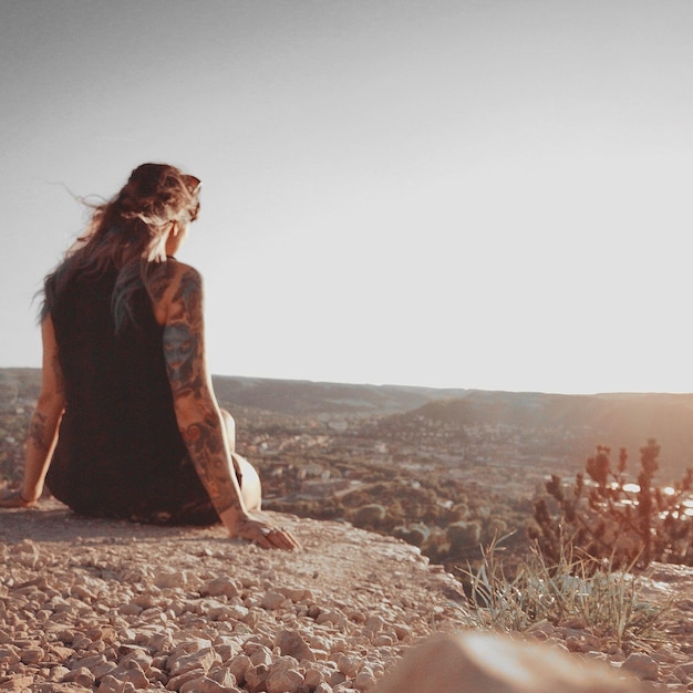 Foto vista traseira de uma mulher olhando para a paisagem enquanto estava sentada em uma rocha contra um céu claro