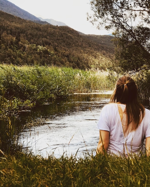 Vista traseira de uma mulher sentada ao lado de um lago contra uma montanha