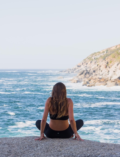 Vista traseira de uma mulher sentada em uma rocha ao lado do mar contra um céu claro