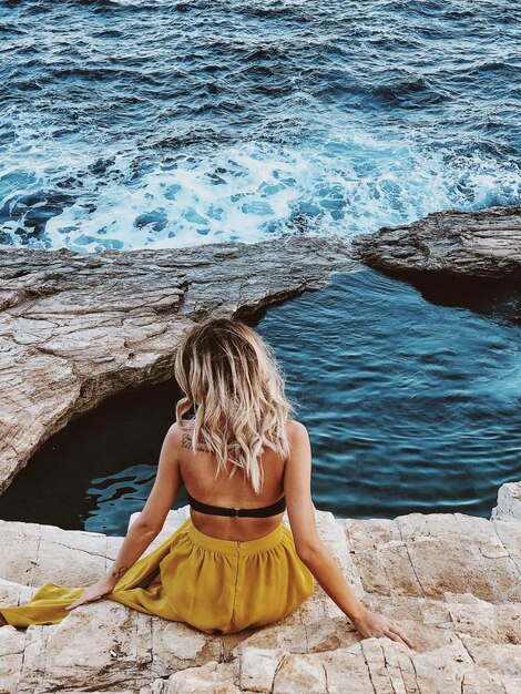 Foto vista traseira de uma mulher sentada em uma rocha ao lado do mar