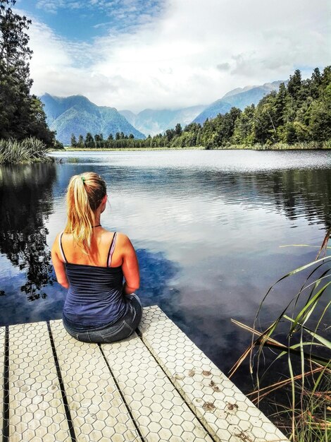 Foto vista traseira de uma mulher sentada no cais sobre o lago contra o céu