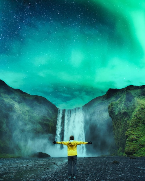 Foto vista traseira de uma turista de jaqueta amarela desfrutando da cachoeira skogafoss e da aurora boreal brilhando à noite na islândia