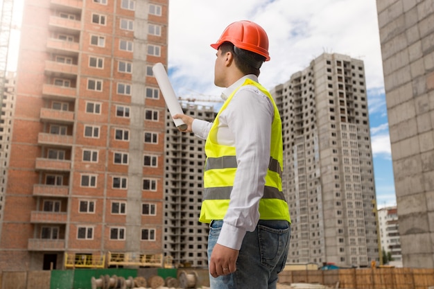 Vista traseira do arquiteto no capacete de segurança apontando com plantas laminadas para o prédio em construção