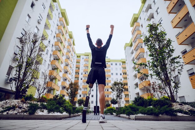 Foto vista traseira do bonito desportista com perna artificial em pé com as mãos para o ar ao ar livre rodeado por edifícios.