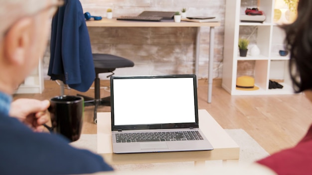 Vista traseira do casal de idosos, sentado no sofá, enquanto acenava para o laptop com tela verde.