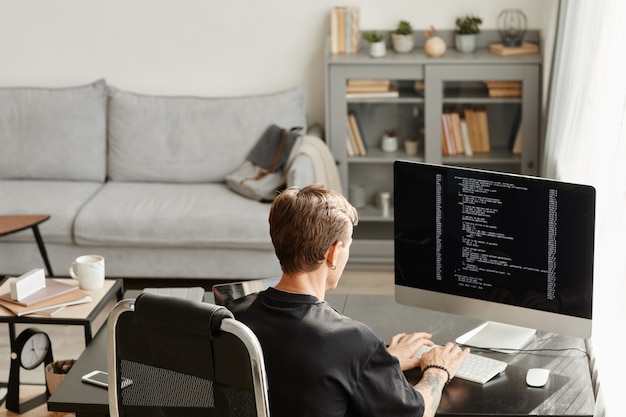 Vista traseira do desenvolvedor de software sentado à mesa em frente ao monitor do computador e desenvolvendo um novo software no escritório