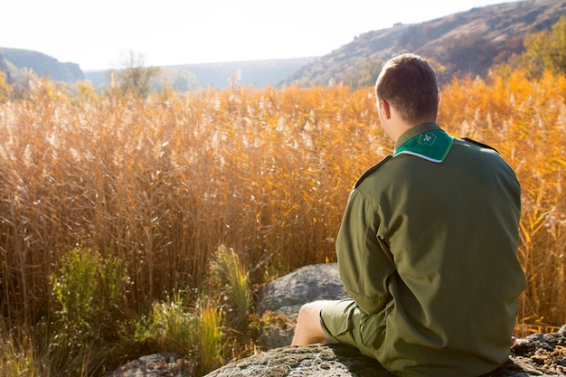 Vista traseira do escoteiro branco sentado na enorme rocha sozinho, observando o amplo campo marrom em uma temporada de outono.