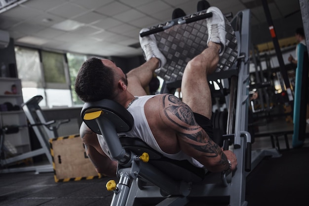 Vista traseira do fisiculturista tatuado irreconhecível malhando na máquina de ginástica leg press