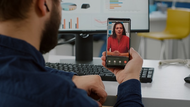 Vista traseira do gerente de projeto segurando smartphone ouvindo empresa líder remota em videochamada, falando online usando fones de ouvido, discutindo em reunião virtual sobre estatística explicativa de projeto financeiro