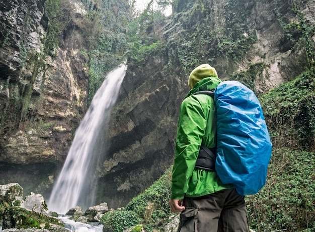 Vista traseira do homem ativo com bolsa de turista com capa de chuva, olhando para a cachoeira