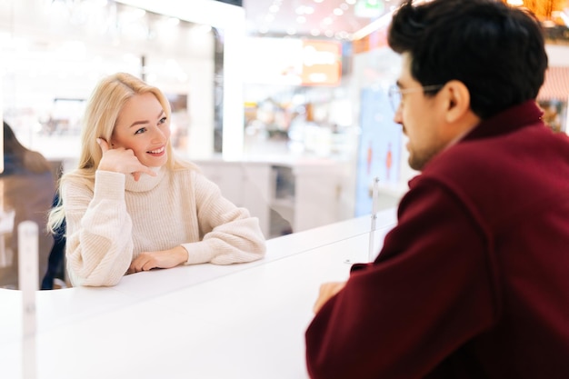 Vista traseira do homem bonito falando conversando através da divisória de vidro com uma linda mulher loira parada no corredor do shopping com interior brilhante