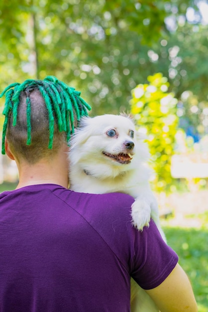 Vista traseira do homem com dreadlocks verdes segurando cachorro branco fofo na rua Spitz mestiço olhando ao redor descansando no ombro do proprietário