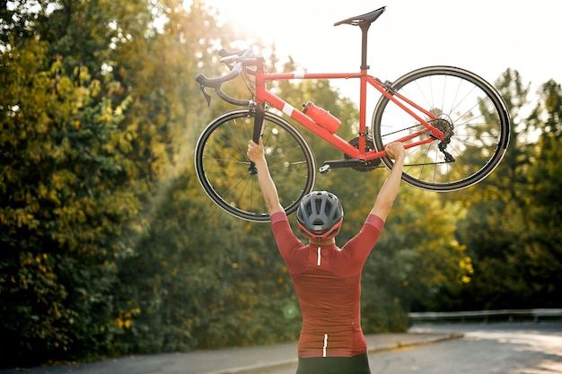 Vista traseira do homem desportivo em roupas esportivas, capacete protetor e óculos carregando a bicicleta...