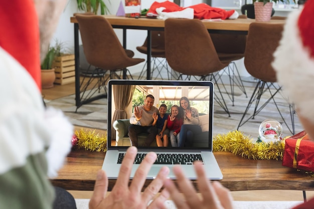 Foto vista traseira do homem e do filho com chapéus de papai noel acenando enquanto faz uma videochamada com a família com chapéus de papai noel wav