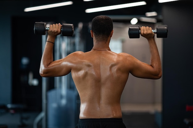 Vista traseira do homem negro atlético com torso nu exercitando com halteres