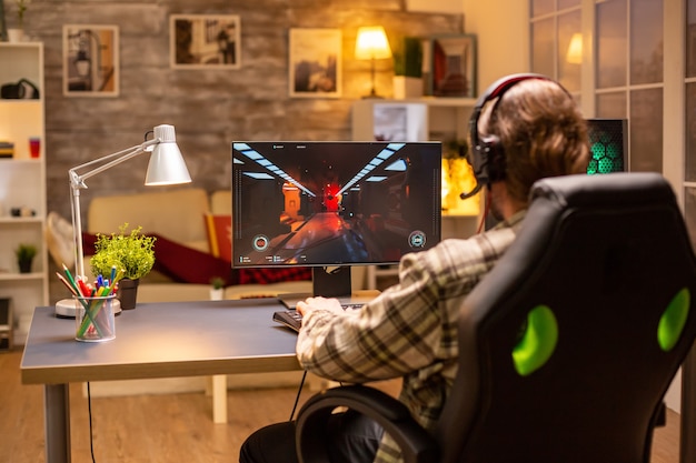 Vista traseira do jogador masculino jogando um atirador em seu poderoso computador PC tarde da noite na sala de estar.