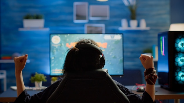 Vista traseira do jogo de tiro espacial ganhando do gamer mulher feliz na sala de casa de jogos. Cyber desempenho em jogos de vídeo de streaming de computador RGB poderosos usando fones de ouvido profissionais para campeonatos online