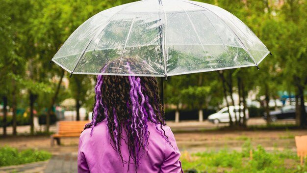 Vista traseira do jovem com guarda-chuva transparente em pé na rua em tempo chuvoso Mulher irreconhecível com dreadlocks encaracolados na camisa roxa na caminhada na chuva