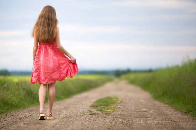 Vista traseira do jovem romântica magro mulher de vestido vermelho com cabelos longos, andando pela estrada de terra ao longo do campo verde num dia ensolarado de verão no céu azul