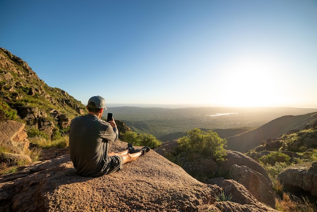 Vista traseira do jovem sentado no topo da montanha tirando foto com smartphone na hora do pôr do sol