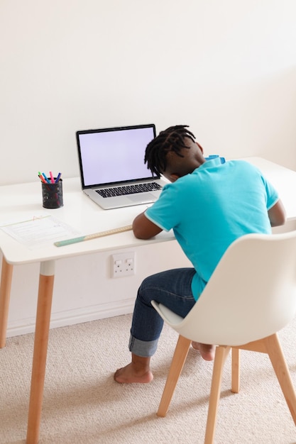 Vista traseira do menino afro-americano com sentado na mesa com o laptop fazendo trabalhos escolares