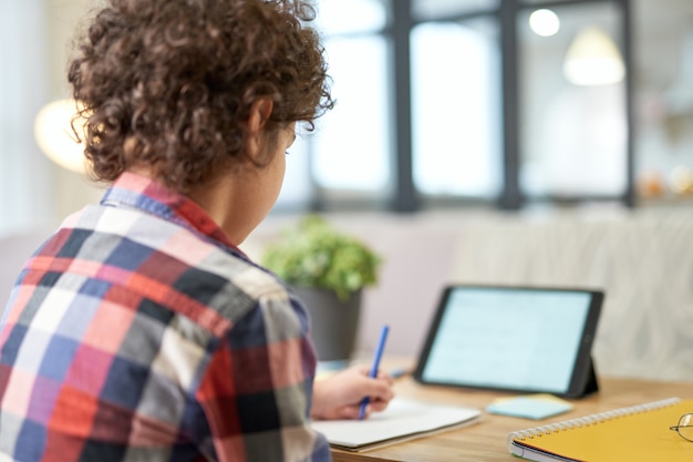 Vista traseira do menino da escola latina fazendo anotações, tendo aula online usando tablet digital enquanto estudava em casa. Educação online, tecnologia, educação domiciliar. Foco seletivo