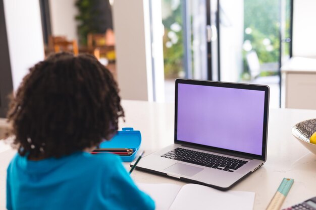 Vista traseira do menino hispânico assistindo aula online através do laptop enquanto está sentado na mesa de jantar