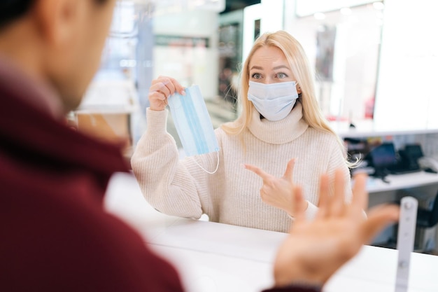 Vista traseira do ombro de um homem confuso irreconhecível falando através de uma divisória de vidro com uma equipe feminina loira em máscara facial apontando para máscara de proteção médica