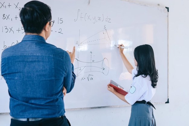 Vista traseira do professor asiático do ensino médio observando o aluno escrevendo no quadro branco na sala de aula