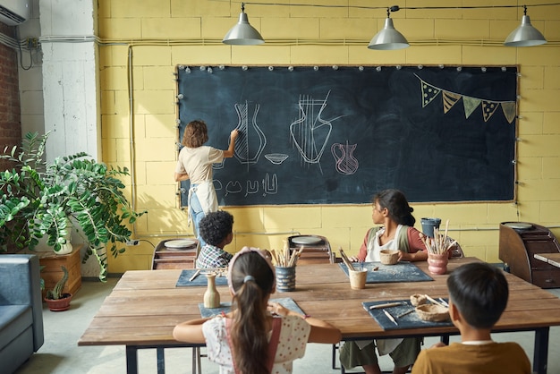 Vista traseira do professor de cerâmica desenhando no quadro-negro enquanto explica a crianças multiétnicas como fazer ...