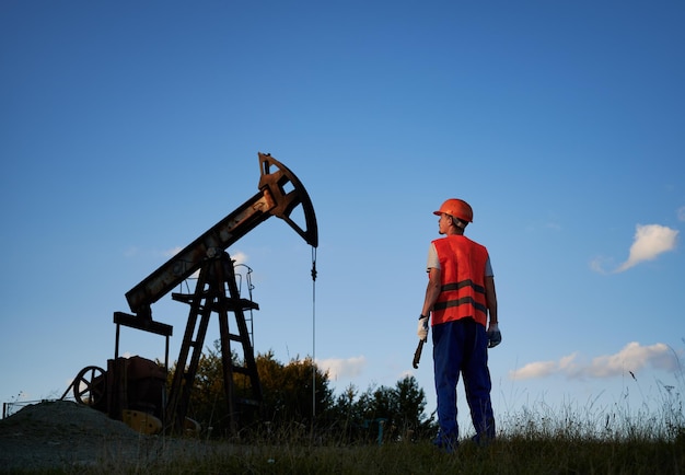 Vista traseira do trabalhador do sexo masculino no capacete protetor em pé perto de jack de bomba de petróleo sob céu azul Engenheiro de petróleo em colete de trabalho segurando chave industrial e olhando para bomba de óleo rockermachine